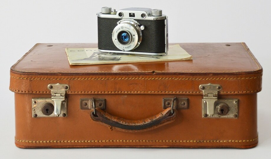 The picture is part of the travel blog 'The Essence of Traveling'. This travel blog features authentic and unique travel content such as compact travel guides and itineraries, travel tips and how-to guides, travel essays, and authentic perspectives about destinations from all around the world. The picture shows an antique brown suitcase in front of a white background. On top of the suitcase, there is an old camera and a magazine. The photo is used for the blog article 'The Ultimate Travel Packing Checklist'.